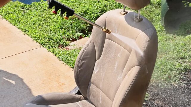 Worker uses pressure washer to clean car seat outside in driveway seat is wet dirt is washing away.