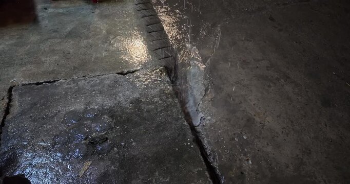 Close up of rainwater falling steadily into a drainage gutter on a cracked concrete street at night during a rain, where water flows into the sewer.