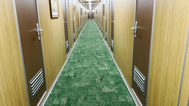 A long corridor with a green carpet stretches inside a cruise ship. The neat wooden doors create a tidy space. The quiet hallway brings a peaceful and calm feeling.