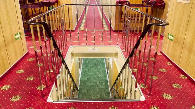 A staircase with metal railings leads down between red and green carpeted hallways inside a cruise ship. The neat interior feels welcoming and cozy. The quiet space gives a calm mood.
