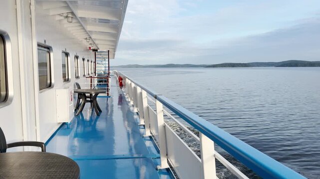 Tables and chairs line the promenade deck as a cruise ship moves across Lake Ladoga. Distant shores sit under a soft bright sky. The quiet breeze creates a peaceful and relaxing mood.