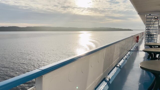 Sunlight glimmers on the water as a cruise ship deck sails across Lake Ladoga. Tables line the quiet railing. The warm atmosphere brings a peaceful and serene feeling.