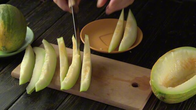 Hands slice ripe melon wedges and arrange them on a plate beside a wooden cutting board, showing fresh fruit preparation on a rustic dark table