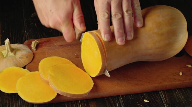 Hands slicing orange butternut squash into round pieces with a kitchen knife on a wooden cutting board, overhead food preparation scene in a rustic kitchen