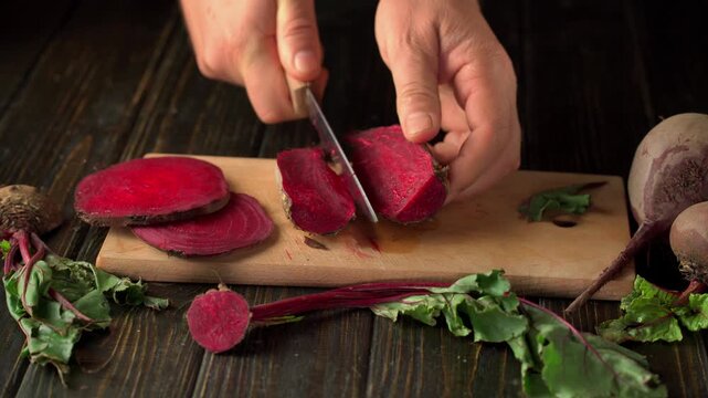 Hands slicing fresh red beetroot with kitchen knife on wooden cutting board, revealing vibrant interior and arranging thin round pieces in rustic kitchen setting