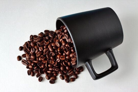 Mainz, Rhineland Palatinate, Germany: Roasted coffee beans spilling from black ceramic mug on clean white background.