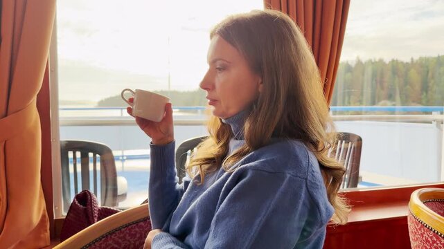 A woman drinks tea while looking out a cruise ship window at Lake Ladoga. Warm light illuminates the quiet room. The calm view fills her with peaceful joy and comfort.
