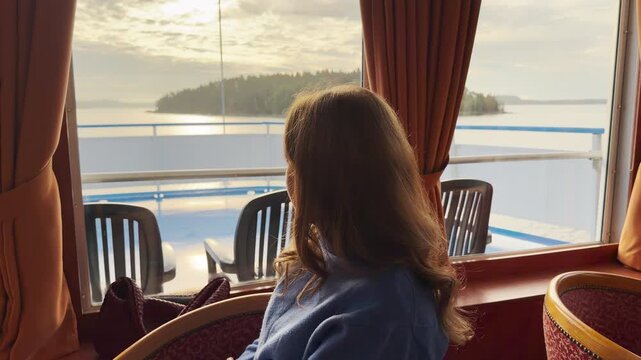 A passenger looks out a large window on a cruise ship as it sails on Lake Ladoga. The soft sunlight shines over distant islands. The gentle scenery brings a peaceful and calm feeling.