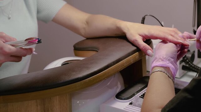 A horizontal video showing a manicurist using a wooden stick on cuticles while the client holds a color palette to choose a gel polish in a salon.