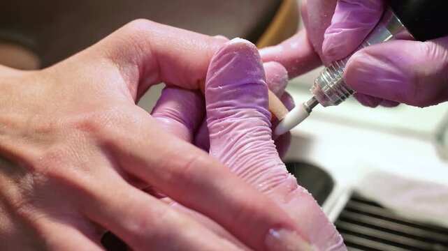 A horizontal macro video showing a manicurist in pink gloves filing and shaping a client's fingernail after applying the first base layer of gel in a salon.