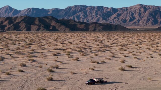 A woman and man travel in an SUV with a teardrop camper trailer through Mojave National Preserve during sunrise among Joshua trees.