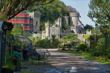 An old stone castle with turrets and a Union Jack flag overlooks a path and foreground garden center structures - Gwrych Castle, Abergele, Wales, UK © Robert