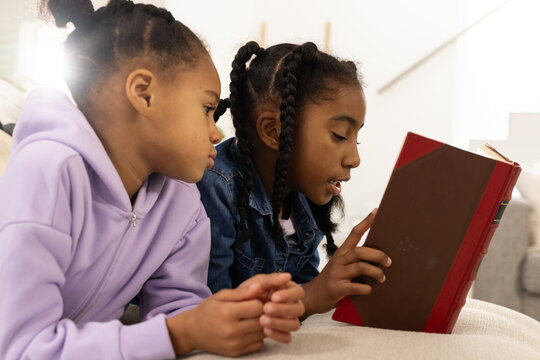 African American school-age girls lying on light sofa reading aloud from red hardcover book