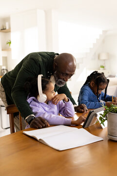 African mature man with two female youths leaning over wooden table at home guiding with tablet