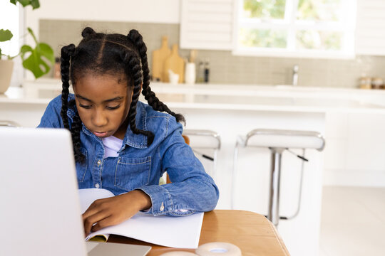 Teen African American girl studying at wooden kitchen table in denim jacket, using laptop, notebook