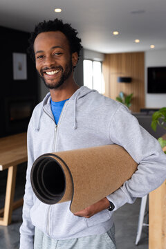 African American man wearing gray hoodie standing at home holding rolled cork mat by dining table