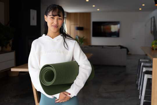 African American woman holding rolled green yoga mat in open-plan home wearing hoodie