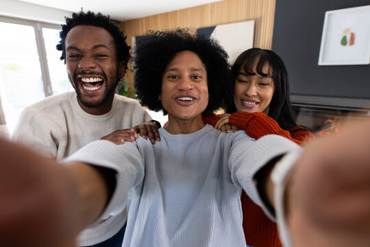 Diverse friends posing for handheld selfie in living room with smartphone, knit sweaters, window