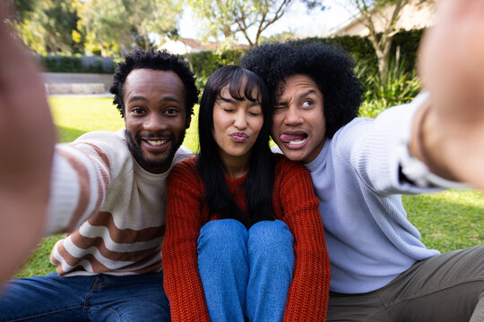 Diverse friends posing for selfie on grassy lawn holding smartphone wearing knit sweaters jeans