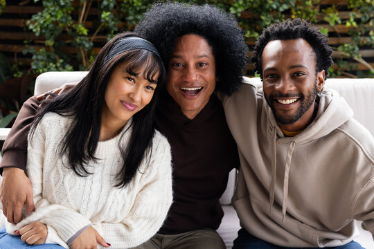 Diverse friends sitting and smiling on light-colored outdoor sofa on planted patio wearing hoodies