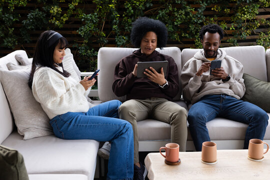 Diverse friends in hoodies sitting on outdoor sectional sofa in backyard using phones, tablet, mugs