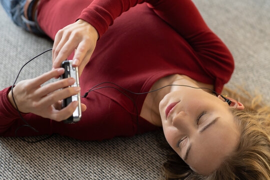 Woman lying on gray carpet at home wearing red top, holding device with wired earbuds, listening