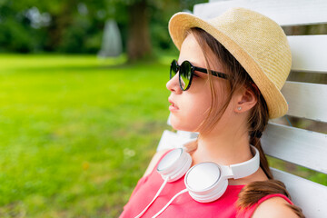 Profile portrait of a teenage girl wearing a straw hat, sunglasses, and white headphones around her neck, relaxing on a white wooden chair in a green park. © bubutu