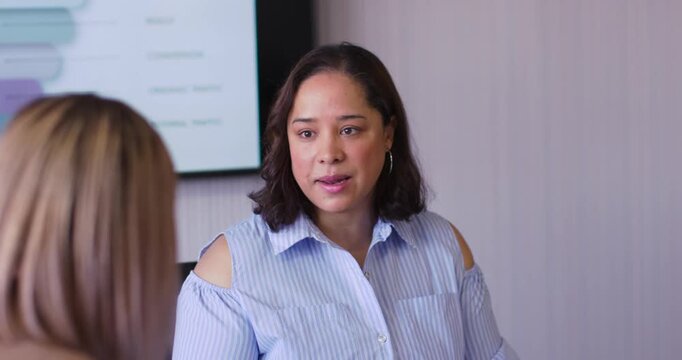 African American woman prompted by wall chart speaking to blonde peer at meeting, explaining data