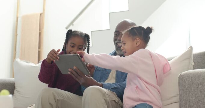 African American family laughing with girls tapping tablet, entertaining man on couch