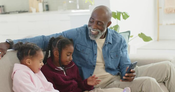Grandpa and grandkids entertaining while grandpa tapping phone and them tapping tablet on sofa