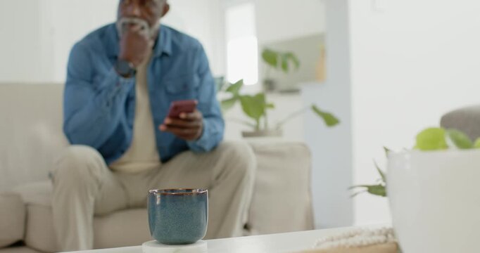 African American man in denim shirt reading phone message smiling sitting on couch near blue mug