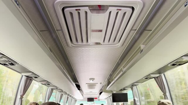 A roof vent circulates air along the ceiling of a moving bus. Passengers travel smoothly past bright green trees. The clean cabin feels calm and peaceful.