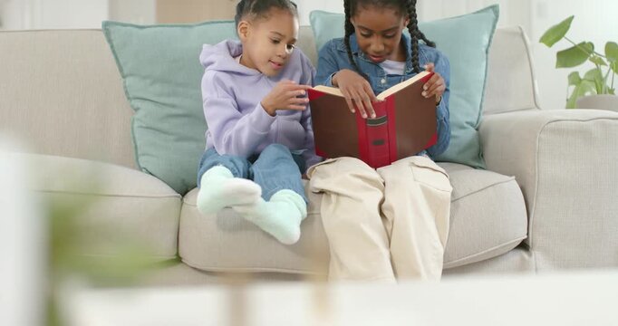 African American girls reading red book on sofa, younger asking question and reader turning pages