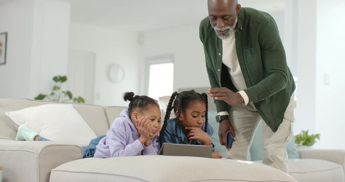 African American family, senior entering with phone guiding two girls using tablet on sofa at home