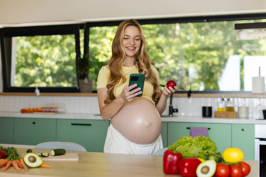 Smiling pregnant woman shopping online on smartphone while holding fresh nectarine in bright kitchen, copy space