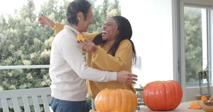 Senior Asian man and African woman on porch, woman holding pulp teasing hugging carving pumpkins