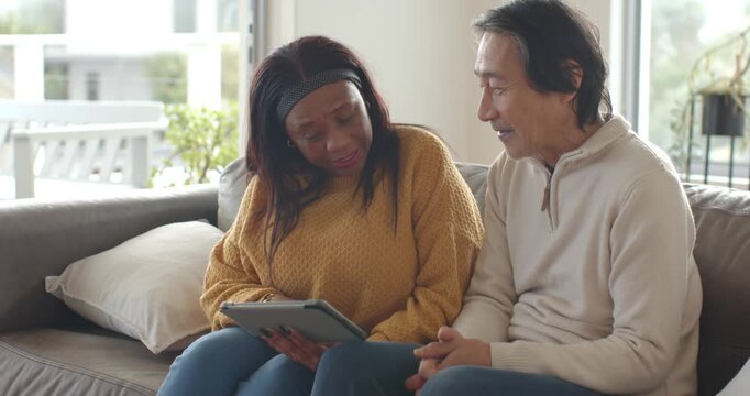 Diverse couple sitting on sofa by window, woman showing tablet, discussing plans while man leans in