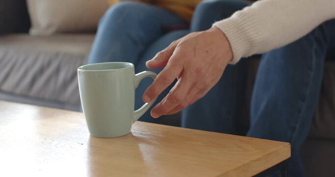 Diverse couple on sofa, senior man reaching for mint-green mug, woman in yellow sweater sipping