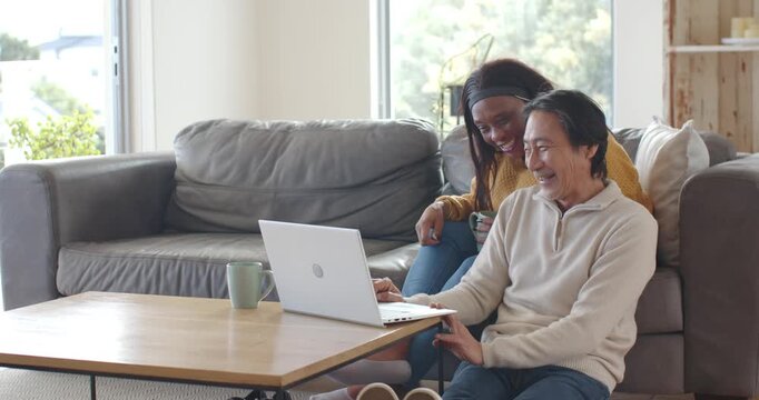 Diverse couple, senior man and woman leaning at home, woman pointing at laptop, man typing, viewing