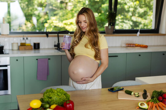 Happy pregnant woman holding glass of milk while gently cradling her baby bump in bright modern kitchen