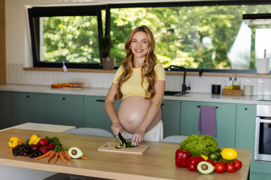 Happy pregnant woman smiling at camera while cutting cucumber preparing fresh vegetables for lunch while standing behind kitchen island