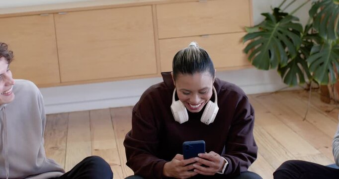 Diverse friends watching woman scrolling phone after ping laughing while leaning on wood floor