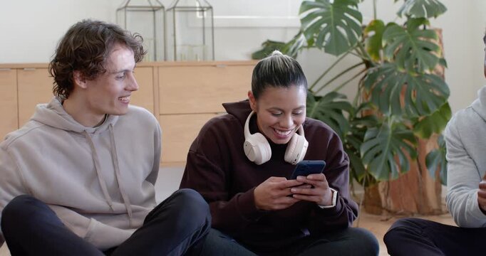 Friends sitting on hardwood floor in living room after man pointing, woman scrolling phone laughing