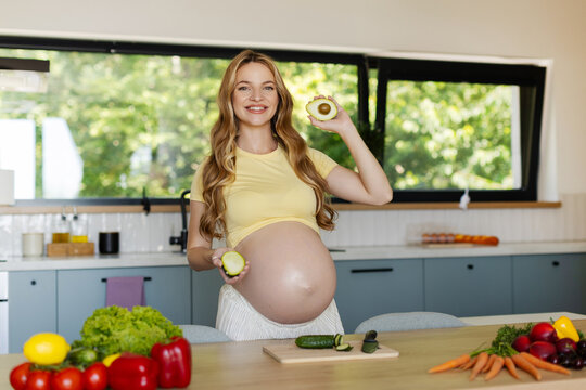 Happy expectant mother showing fresh avocados while preparing healthy meal with colorful vegetables in contemporary home kitchen