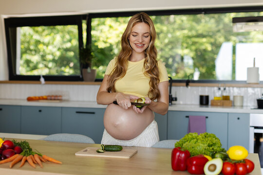 Smiling pregnant woman cutting ripe avocado in modern kitchen while preparing healthy meal with fresh vegetables, free space