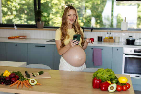 Cheerful pregnant woman texting on her smartphone while holding fresh nectarine, standing in bright modern kitchen