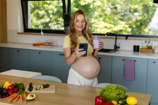 Happy expectant mother enjoying glass of milk while reading healthy pregnancy article on her smartphone in contemporary kitchen
