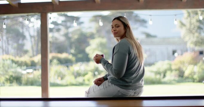 Adult woman sitting on windowsill under pergola pausing with mug and savoring morning light in yard