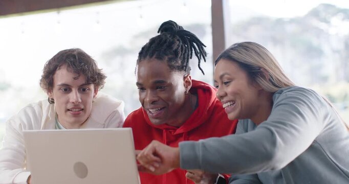 Diverse friends in hoodies leaning over silver laptop at cafe, pointing, clapping upon seeing win