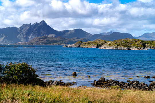 Autumn coastal landscape near Mortsund in the Lofoten Islands with blue sea, rocky islands, rugged mountains and a cloudy blue sky.